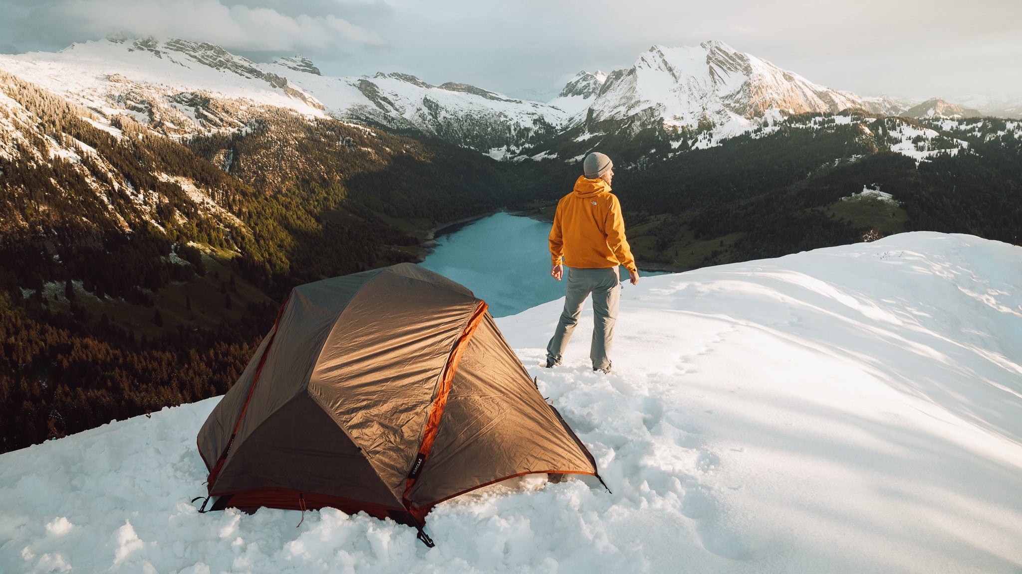 Person standing on a snowy mountain with a tent nearby, surrounded by snow-covered mountains.